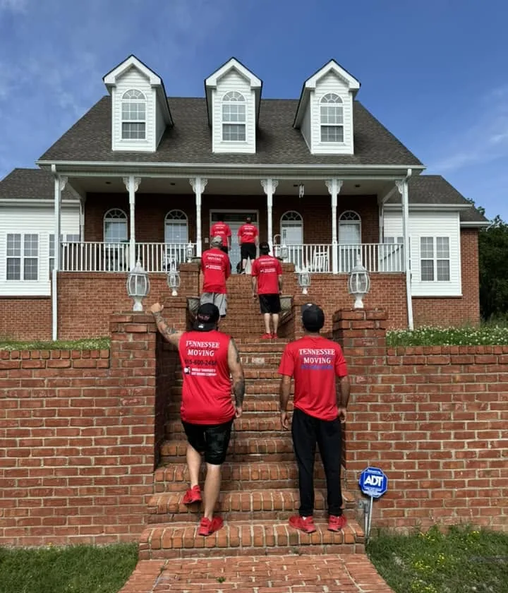 Members of the moving crew in red Tennessee Country Boys Moving shirts walking up the brick front steps of a two-story house.