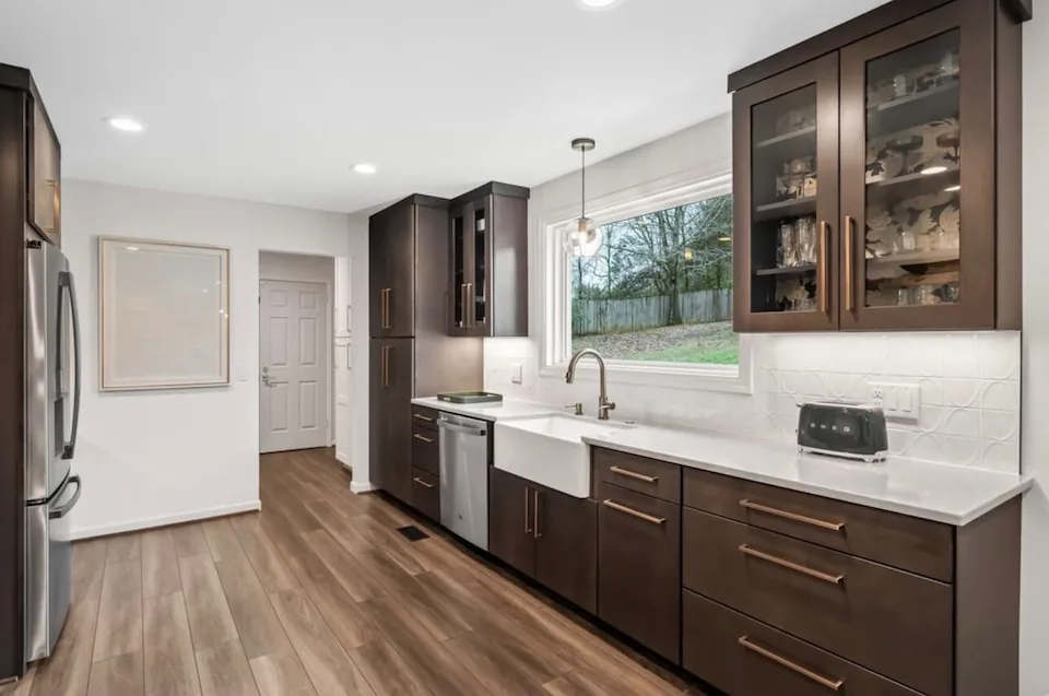 A kitchen with dark cabinets, a farmhouse sink under a window and wood-look flooring.