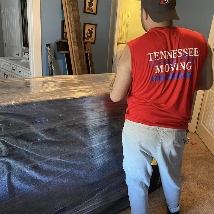 A mover in a red Tennessee Country Boys Moving shirt beside a piece of furniture padded and shrink-wrapped for the move.