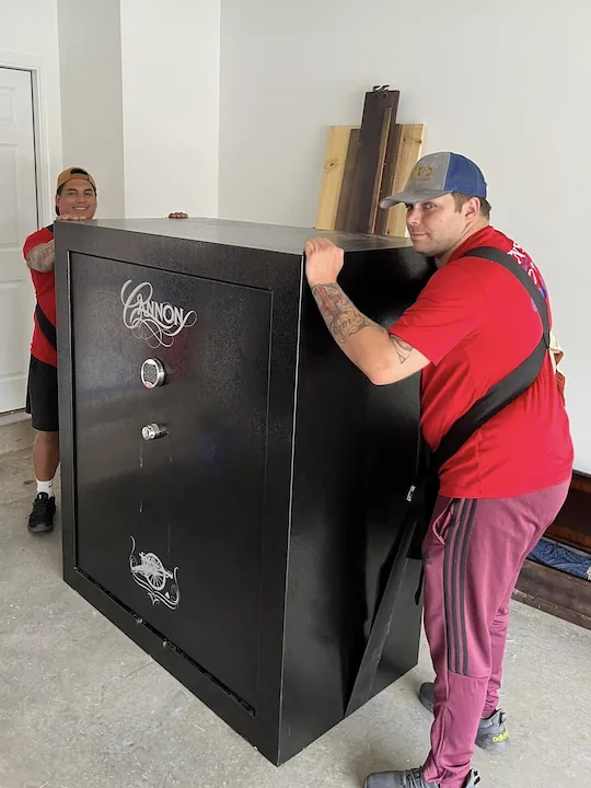 Two movers in red shirts using lifting straps to move a large gun safe.